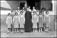 Retrato de grupo de meninas e um padre, junto a um portão, em local não identificado